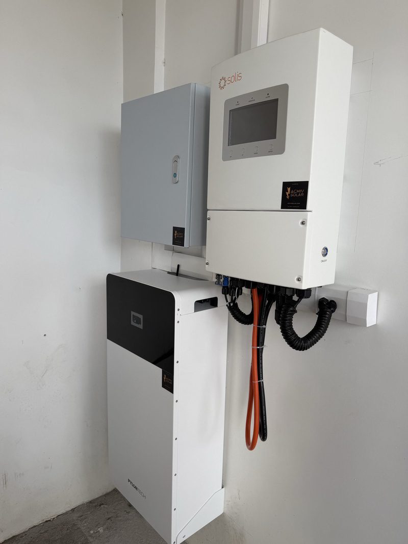 Close view of the hybrid inverter, protection box and battery, cables bundled in black conduit.