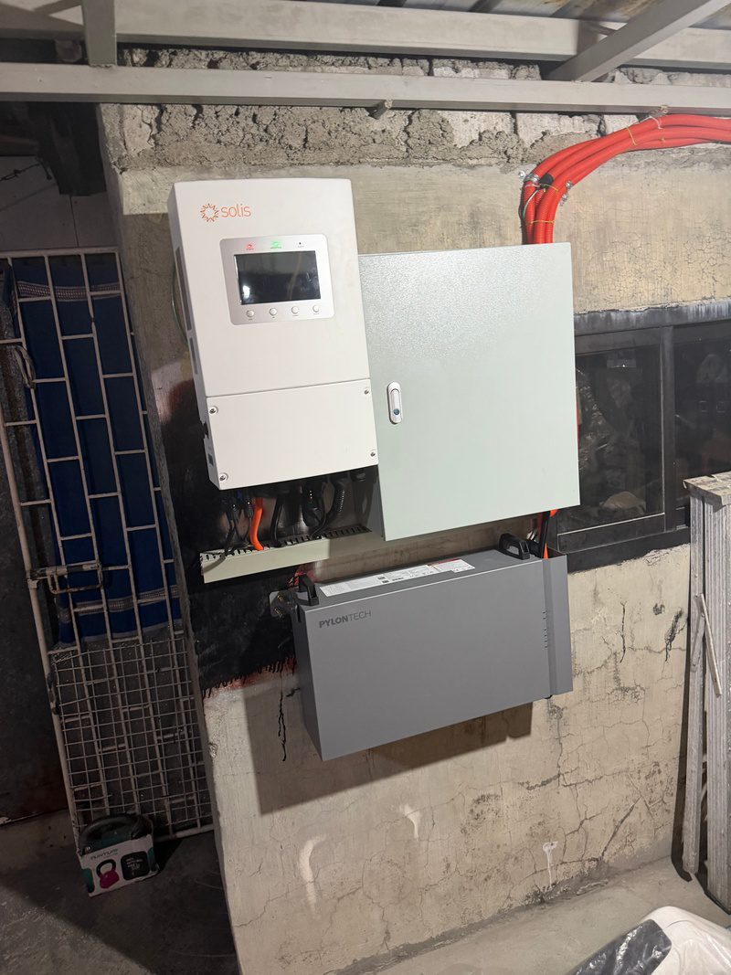 The finished equipment wall from the side: inverter, protection box and battery, with the orange conduit sweeping up to the roof.