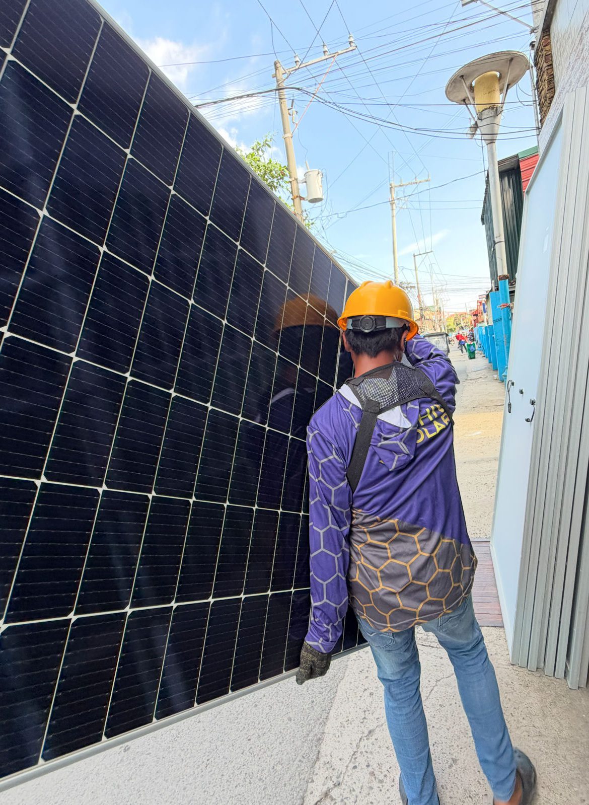 An Achiv installer in a hard hat, seen from behind, walking a full-size solar panel down a narrow Metro Manila street to the job.
